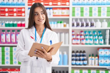 Young beautiful hispanic woman pharmacist smiling confident writing on notebook at pharmacy