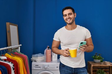 Genç İspanyol adam akıllı telefon kullanıyor ve kahve içiyor çamaşır odasında çamaşır makinesini bekliyor.