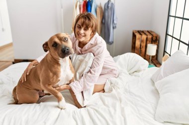 Beyaz kadın gülümsüyor. Köpeğiyle oynuyor. Yatak odasında oturuyor.