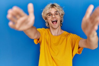 Young modern man standing over blue background looking at the camera smiling with open arms for hug. cheerful expression embracing happiness. 