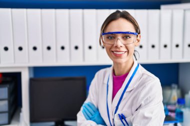 Genç ve güzel İspanyol bilim kadını gülümsüyor. Kollarını kavuşturup laboratuvarda oturuyor.