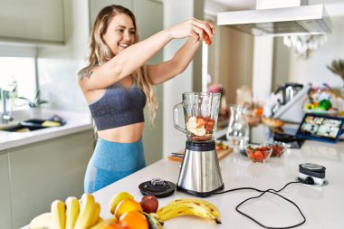 Young woman smiling confident pouring strawberries on blender at kitchen