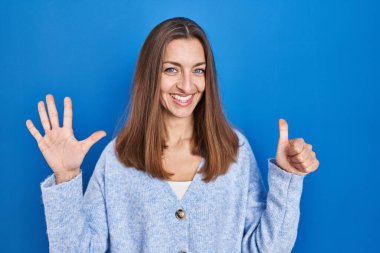 Young woman standing over blue background showing and pointing up with fingers number six while smiling confident and happy. 