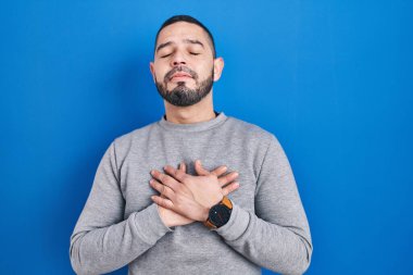 Mavi arka planda duran İspanyol adam elleri göğsünde, gözleri kapalı ve yüzünde minnet dolu bir jest ile gülümsüyor. sağlık kavramı. 