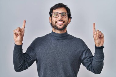 Handsome latin man standing over isolated background smiling amazed and surprised and pointing up with fingers and raised arms. 