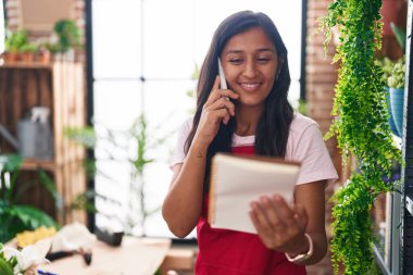 Young beautiful hispanic woman florist talking on smartphone reading notebook at flower shop