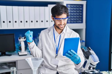 Handsome latin man working at scientist laboratory using tablet screaming proud, celebrating victory and success very excited with raised arm 