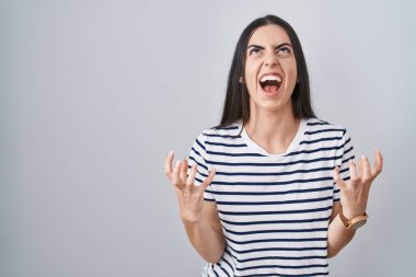 Young brunette woman wearing striped t shirt crazy and mad shouting and yelling with aggressive expression and arms raised. frustration concept. 