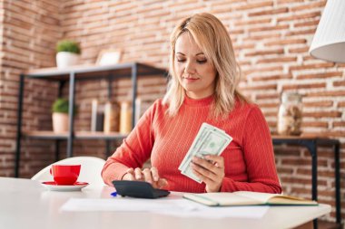 Young blonde woman sitting on table counting dollars at home