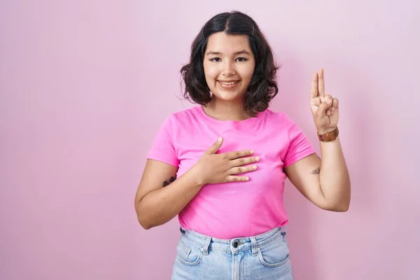 ピンクの背景に立っている若いヒスパニック系の女性は 胸や指を手で身に着けて笑顔 忠誠の誓いを作ります — ストック写真
