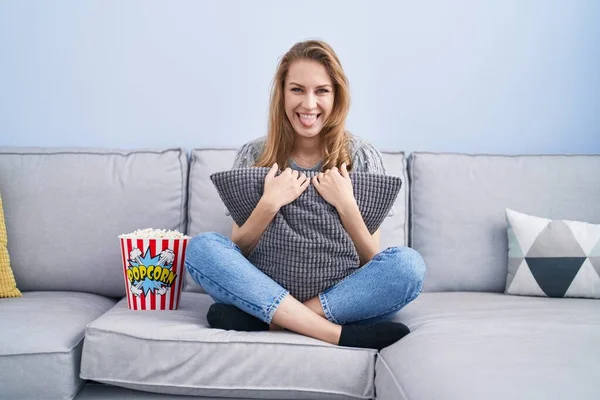Beautiful blonde woman watching tv sitting on the sofa sticking tongue out happy with funny expression.