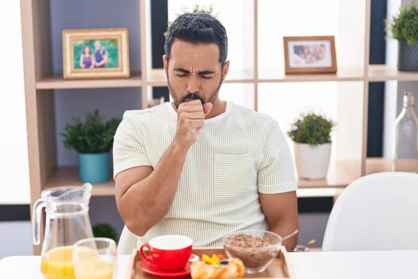 Hispanic man with beard eating breakfast feeling unwell and coughing as symptom for cold or bronchitis. health care concept.