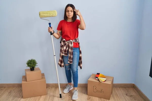 Young hispanic woman painting home walls with paint roller shooting and killing oneself pointing hand and fingers to head like gun, suicide gesture.