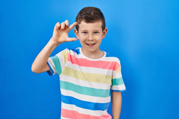 青い背景の上に立つ若い白人の子供は笑顔で 指を見てカメラで小さなサイズのサインをする手で自信を持ってジェスチャー 概念を測定し — ストック写真