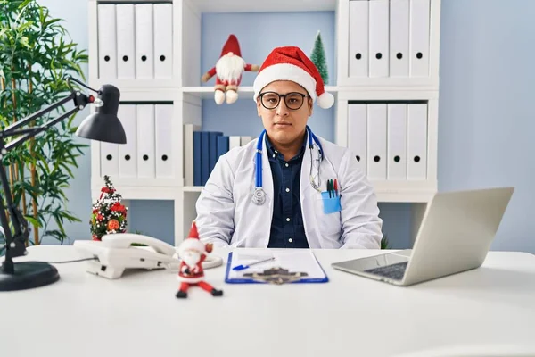 Hispanic young doctor man at the clinic on christmas with serious expression on face. simple and natural looking at the camera.