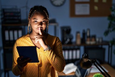 Saçları örgülü Afro-Amerikalı bir kadın geceleri ofiste çalışıyor tabletle dudaklarında parmaklarıyla sessiz olmak istiyor. sessizlik ve gizli konsept. 