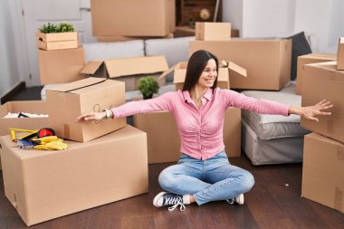 Genç, güzel, İspanyol bir kadın yeni evinde yerde oturmuş kollarını açıyor.