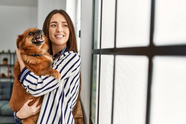 Genç İspanyol kadın gülümsüyor. Kendine güvenen, sarılan köpek evde duruyor.