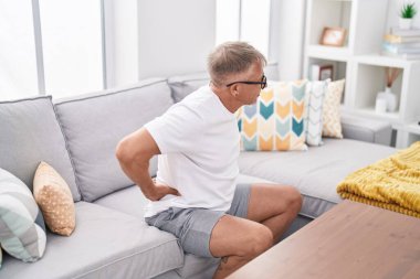 Orta yaşlı, kır saçlı, sırt ağrısı çeken bir adam evdeki kanepede oturuyor.
