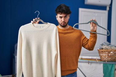 Hispanic man with beard holding sweater on hanger at laundry room skeptic and nervous, frowning upset because of problem. negative person. 