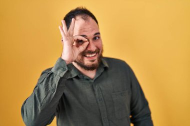 Plus size hispanic man with beard standing over yellow background doing ok gesture with hand smiling, eye looking through fingers with happy face. 