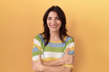 Orta yaşlı İspanyol kadın, sarı arka planda durup kollarını kavuşturup kameraya bakarken gülümsüyor. pozitif kişi. 