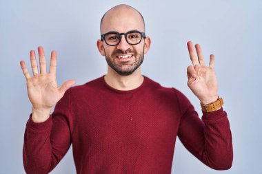 Beyaz arka planda sakallı genç, kel bir adam gözlük takıyor ve 8 numaralı parmaklarıyla işaret ederken kendinden emin ve mutlu bir şekilde gülümsüyor.. 