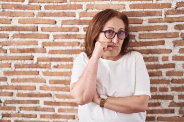 Senior woman with glasses standing over bricks wall mouth and lips shut as zip with fingers. secret and silent, taboo talking 
