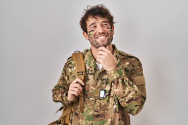 Hispanic young man wearing camouflage army uniform with hand on chin thinking about question, pensive expression. smiling and thoughtful face. doubt concept. 