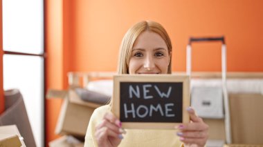 Young blonde woman smiling confident holding blackboard at new home