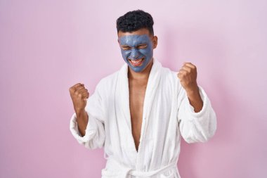 Young hispanic man wearing beauty face mask and bath robe very happy and excited doing winner gesture with arms raised, smiling and screaming for success. celebration concept. 