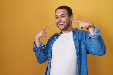 Hispanic man standing over yellow background smiling cheerful showing and pointing with fingers teeth and mouth. dental health concept. 