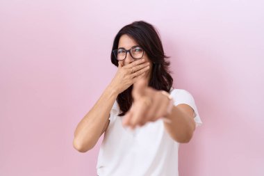 Orta yaşlı, beyaz tişörtlü ve gözlüklü bir İspanyol kadın sana gülüyor, kamerayı işaret ediyor, ağzının üzerinde utanç ifadesi var. 