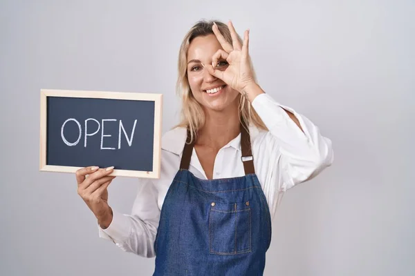 stock image Young blonde woman wearing apron holding blackboard with open word smiling happy doing ok sign with hand on eye looking through fingers 