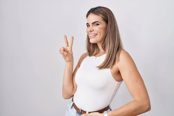 西班牙裔年轻女子站在白色的背景上 微笑着看着摄像机 手指头在做胜利的标志 第二点 — 图库照片