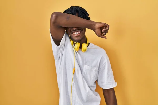 黄色の背景の上に立っているドレッドロックを持つ若いアフリカ人男性は 陽気で面白い腕で目を覆います 視覚障害の概念 — ストック写真