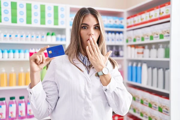 薬局で働いているヒスパニック系の若い女性は 手で口をカバークレジットカードを保持し ショックを受け 間違いを恐れています 驚きの表情 — ストック写真