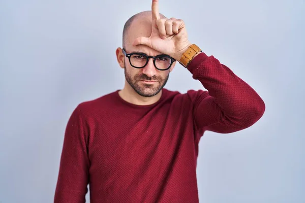 Young bald man with beard standing over white background wearing glasses making fun of people with fingers on forehead doing loser gesture mocking and insulting.