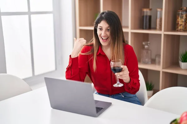 Young brunette woman doing video call drinking red wine pointing thumb up to the side smiling happy with open mouth