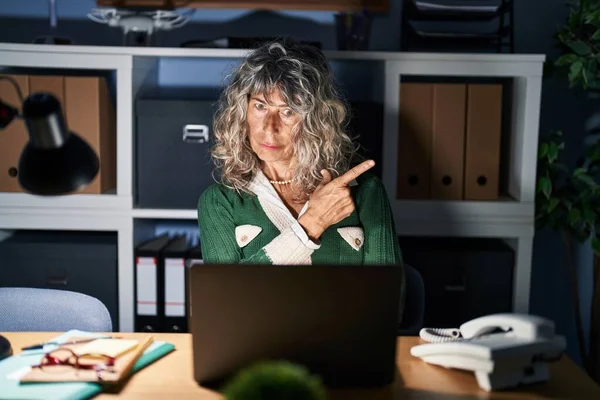Middle age woman working at night using computer laptop pointing with hand finger to the side showing advertisement, serious and calm face