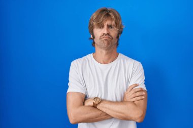 Mavi arka planda duran orta yaşlı bir adam şüpheci ve gergin, yüzünde çapraz kollu tasvip etmeyen bir ifade. negatif kişi. 