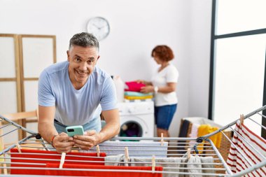 Orta yaşlı erkek ve kadın çamaşırhanede çamaşır asan akıllı telefon kullanan bir çift.