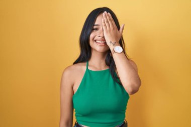 Sarı arka planda duran esmer kadın bir gözünü eliyle kapatıyor, yüzünde kendine güvenen bir gülümseme ve sürpriz bir duygu var.. 