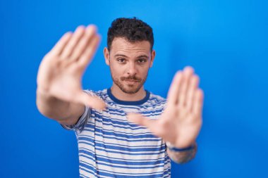 Young hispanic man standing over blue background doing frame using hands palms and fingers, camera perspective 