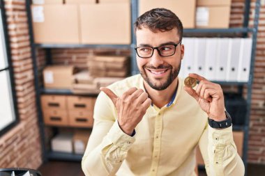 Küçük işletme ecommerce çalışan yakışıklı İspanyol adam Bitcoin 'i elinde tutuyor başparmağıyla yan tarafa bakıyor mutlu bir şekilde ağzı açık gülümsüyor. 