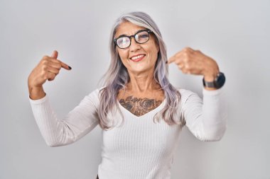 Beyaz arka planda beyaz saçları olan orta yaşlı bir kadın yüzünde bir gülümsemeyle gururla ve mutlu bir şekilde kendini işaret ediyor.. 