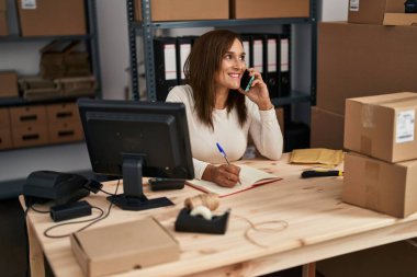 Middle age woman ecommerce business worker talking on the smartphone writing on book at office