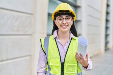 Young hispanic woman architect holding blueprints at street