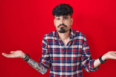 Young hispanic man with beard standing over red background clueless and confused expression with arms and hands raised. doubt concept. 