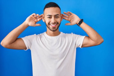 Young hispanic man standing over blue background smiling pulling ears with fingers, funny gesture. audition problem 